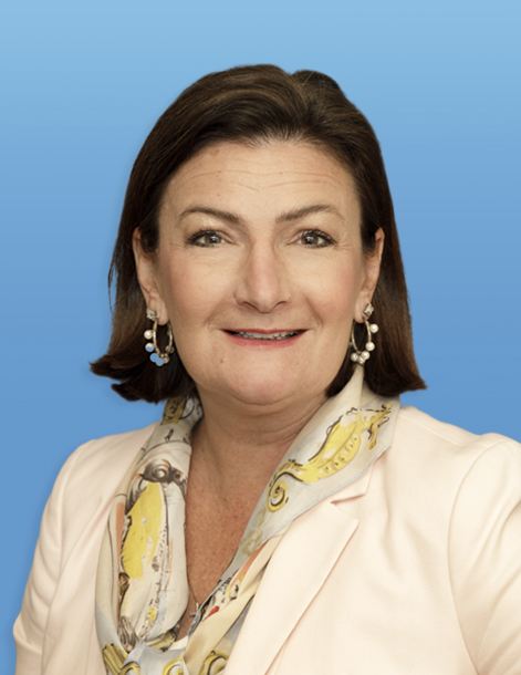 A person with medium-length brown hair is smiling. They are wearing a light-colored blazer and a patterned scarf, with pearl-like earrings, against a solid blue background.