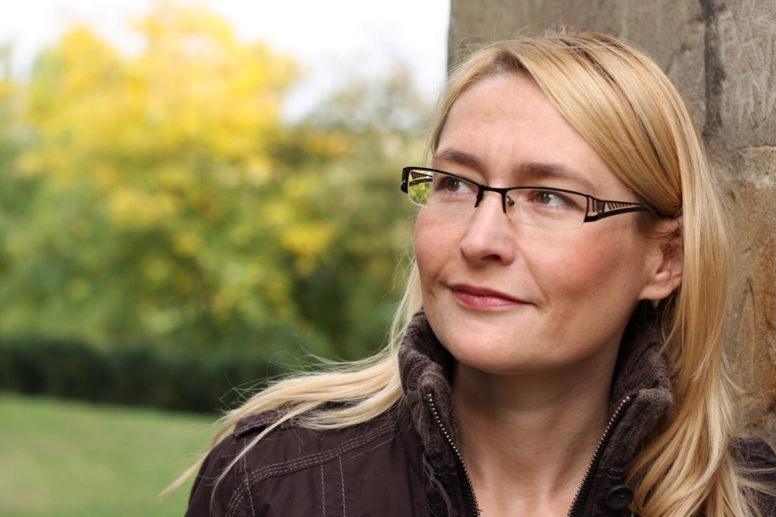 A woman with blonde hair and glasses looks thoughtfully to the side, standing outdoors against a stone wall. She wears a dark jacket, and there are green trees with yellow leaves in the blurred background.
