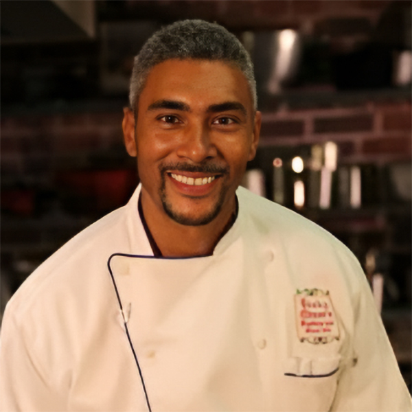 A chef with short grey hair and a beard is smiling in a kitchen. He is wearing a white chefs coat with an embroidered logo. The background features shelves with cooking utensils and a brick wall.