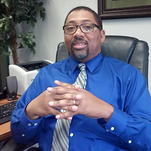 A man wearing glasses, a blue shirt, and a striped tie sits in a leather chair, hands clasped. He is in an office setting with a plant, a printer, and a computer keyboard visible in the background.