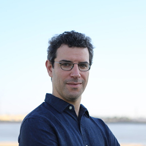 A man with short curly hair and glasses is standing outdoors, wearing a dark button-up shirt. The background is a blurred, light blue sky with a faint horizon.