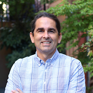 A man with dark hair pulled back is smiling and standing outdoors. He is wearing a light blue plaid shirt. Green leafy trees are in the background.