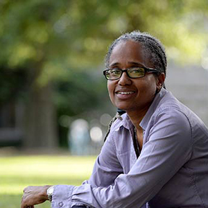A person wearing glasses and a purple shirt sits outdoors on the grass, smiling slightly. The background is blurred, with green foliage indicating a park or garden setting.