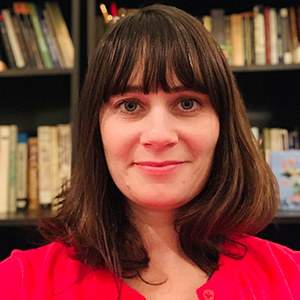 A person with shoulder-length brown hair and bangs is smiling, wearing a red top. They are standing in front of a bookshelf filled with various books.