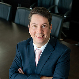 A person in a suit with a pink tie is smiling at the camera, arms crossed, seated at a dark wooden conference table with black chairs in the background.