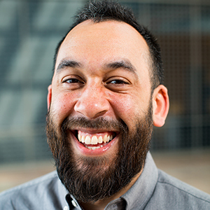 A bearded man smiling widely at the camera, wearing a gray button-up shirt. The background is blurred, suggesting an indoor setting.