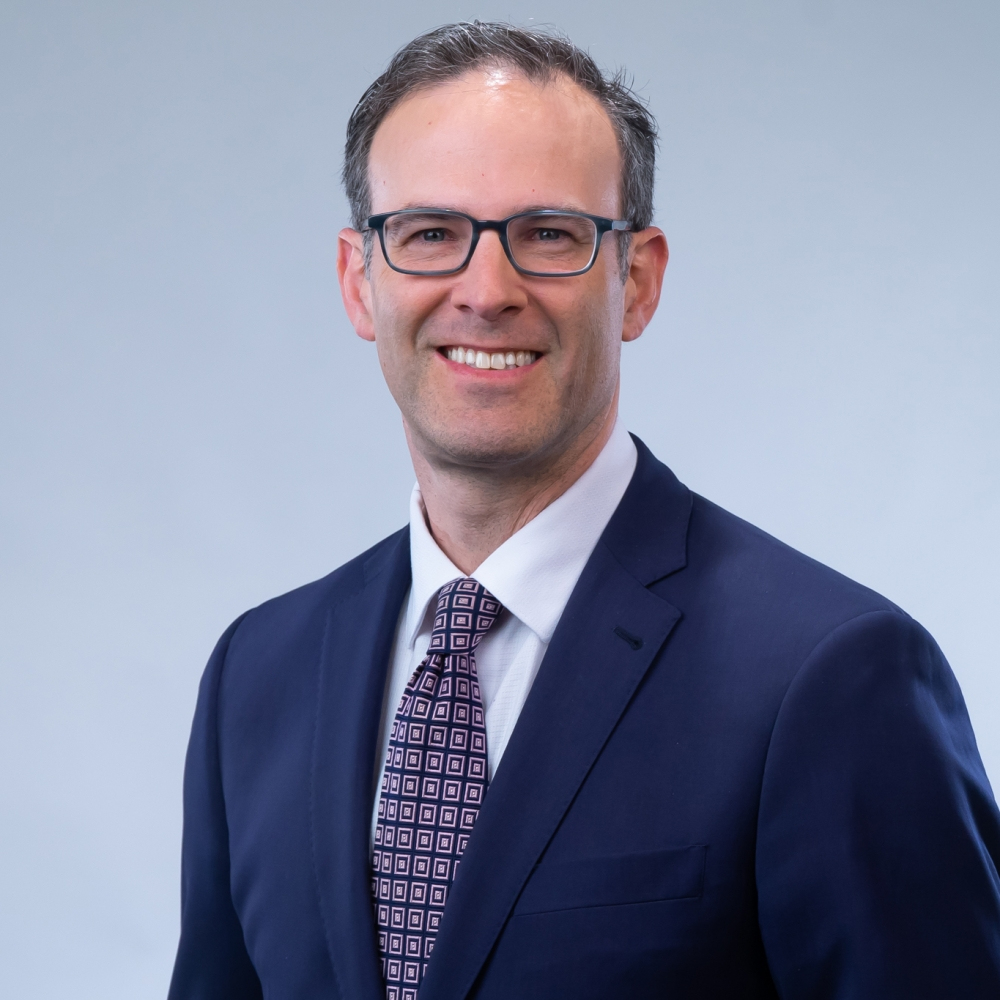A man in a dark blue suit, white shirt, and patterned tie is smiling against a plain light gray background. He is wearing glasses and has short hair.