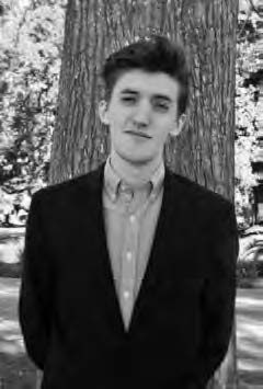A young man wearing a suit stands in front of a large tree. The background shows a park-like setting with trees and sunlight filtering through the leaves. The image is in black and white.