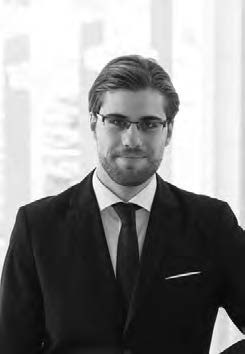 A man in a suit and tie, wearing glasses, stands confidently in front of a blurred background. The image is black and white, showcasing a professional and composed demeanor.