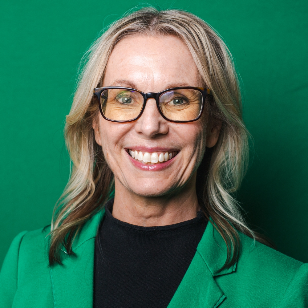 A smiling woman with long blonde hair and eyeglasses is wearing a green blazer over a black shirt, standing against a green background.