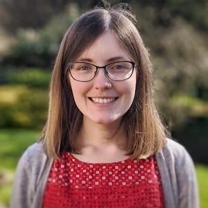A woman with shoulder-length brown hair and glasses is smiling. She is wearing a red patterned top and a gray cardigan. The background is a blurred outdoor setting with greenery.