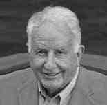 An older man with white hair, wearing a light-colored jacket and shirt, is smiling slightly in a black and white photograph. The background appears dark, creating contrast with the subject.