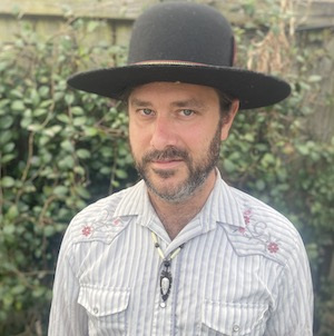 A person with a beard wearing a black wide-brimmed hat and a light-colored, embroidered button-up shirt stands in front of a leafy background.