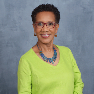 A person with short curly hair, wearing glasses and a yellow-green top, smiles confidently. They accessorize with a blue necklace and earrings against a textured gray background.