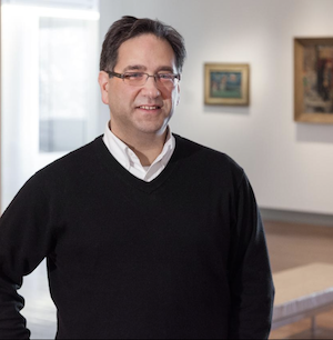 A person with short dark hair, glasses, and wearing a black sweater over a white shirt, stands in an art gallery. Paintings are visible on the wall in the background.