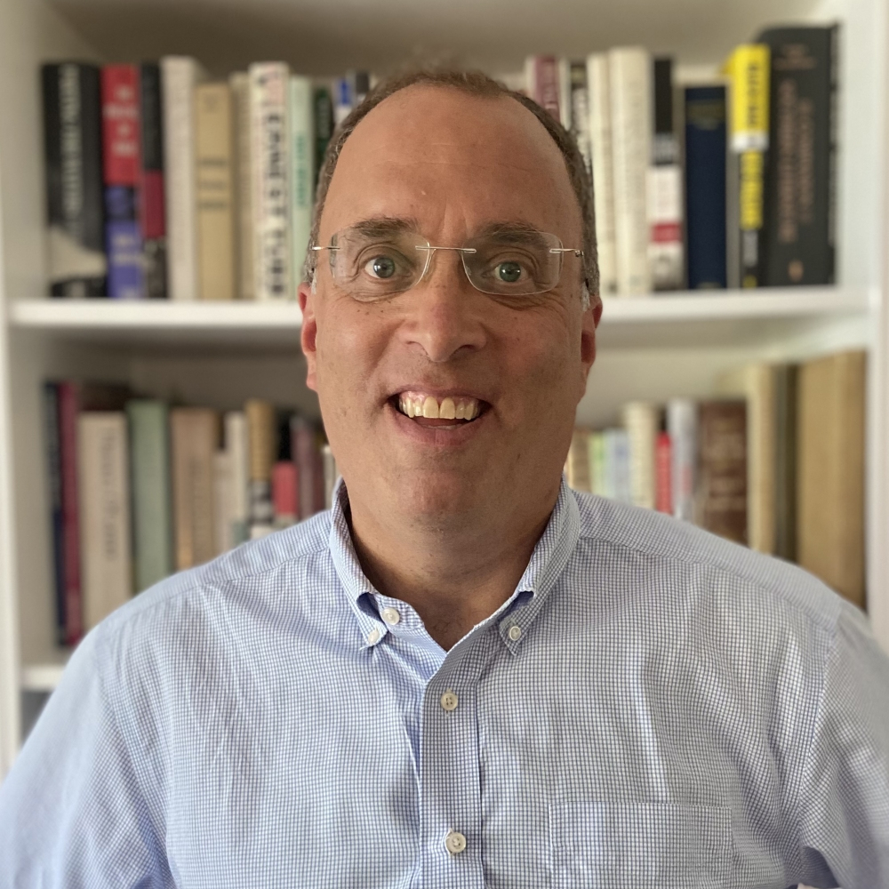 A person wearing glasses and a blue checkered shirt is standing in front of a bookshelf filled with various books. They are smiling at the camera.