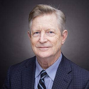 Elderly man with light hair wearing a dark suit, striped tie, and blue shirt, looking at the camera against a dark background.