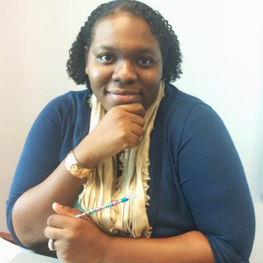 A woman with short curly hair smiles while sitting at a table, resting her chin on her hand. She wears a blue top and a beige scarf, holding a colorful pen in her other hand.