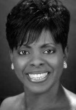 A person smiling, with short dark hair and wearing earrings, set against a dark background. The image is in black and white.