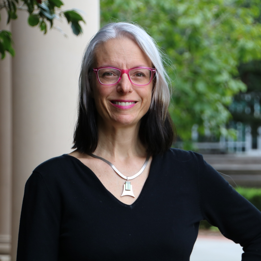 A woman with long, gray-and-black hair, wearing pink glasses and a black top, stands outside. She smiles confidently with her hand on her hip. A green, leafy background and a column are visible behind her.