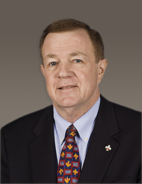 Portrait of a man wearing a dark suit, light blue shirt, and a colorful patterned tie. He has short, light brown hair and is against a plain gray background.