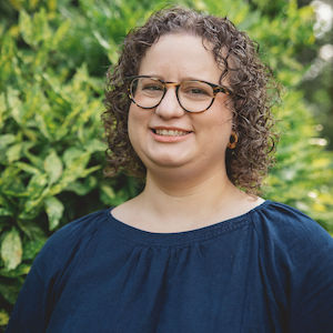 A person with curly hair and glasses smiles while standing in front of green leafy bushes, wearing a dark blue top.