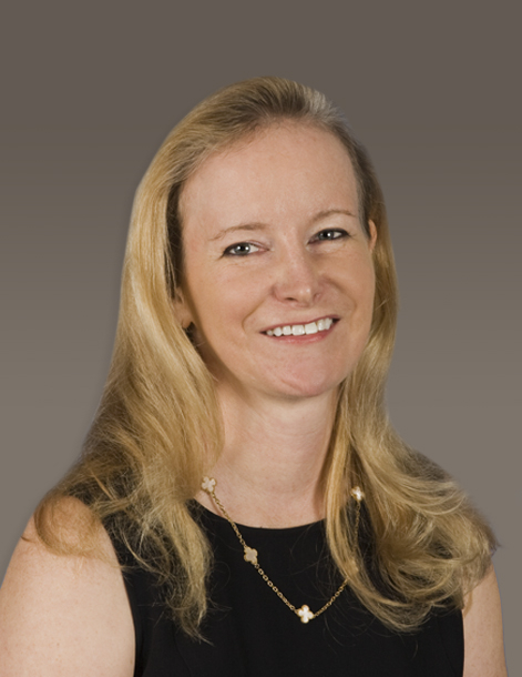Smiling woman with long blond hair wearing a black top and a gold necklace, set against a gray background.