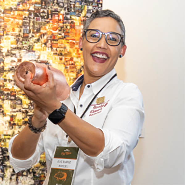 Person smiling and holding a pink bottle, wearing a white shirt with a name tag and black watch. The background features a colorful mosaic or collage.