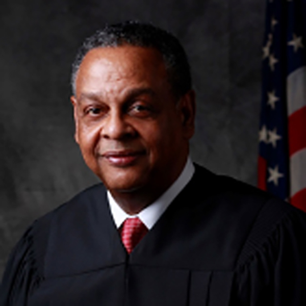 A man in a judges robe stands against a dark background. He wears a white shirt, red tie, and black judicial robe. The U.S. flag is partially visible behind him, suggesting a legal or governmental setting.