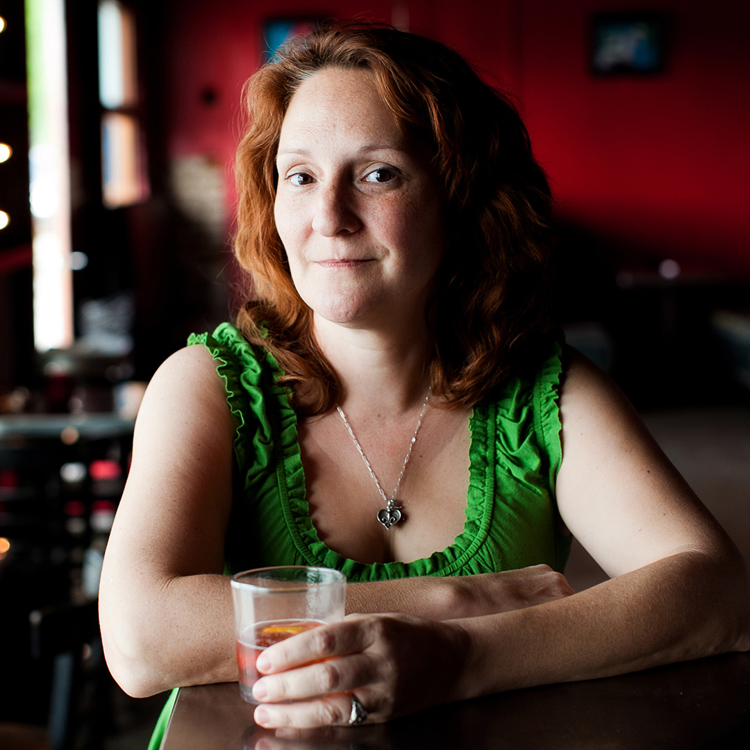 A woman with curly red hair sits at a table in a dimly lit room, wearing a green sleeveless top and a necklace. She holds a glass and smiles softly, with a blurred red background behind her.
