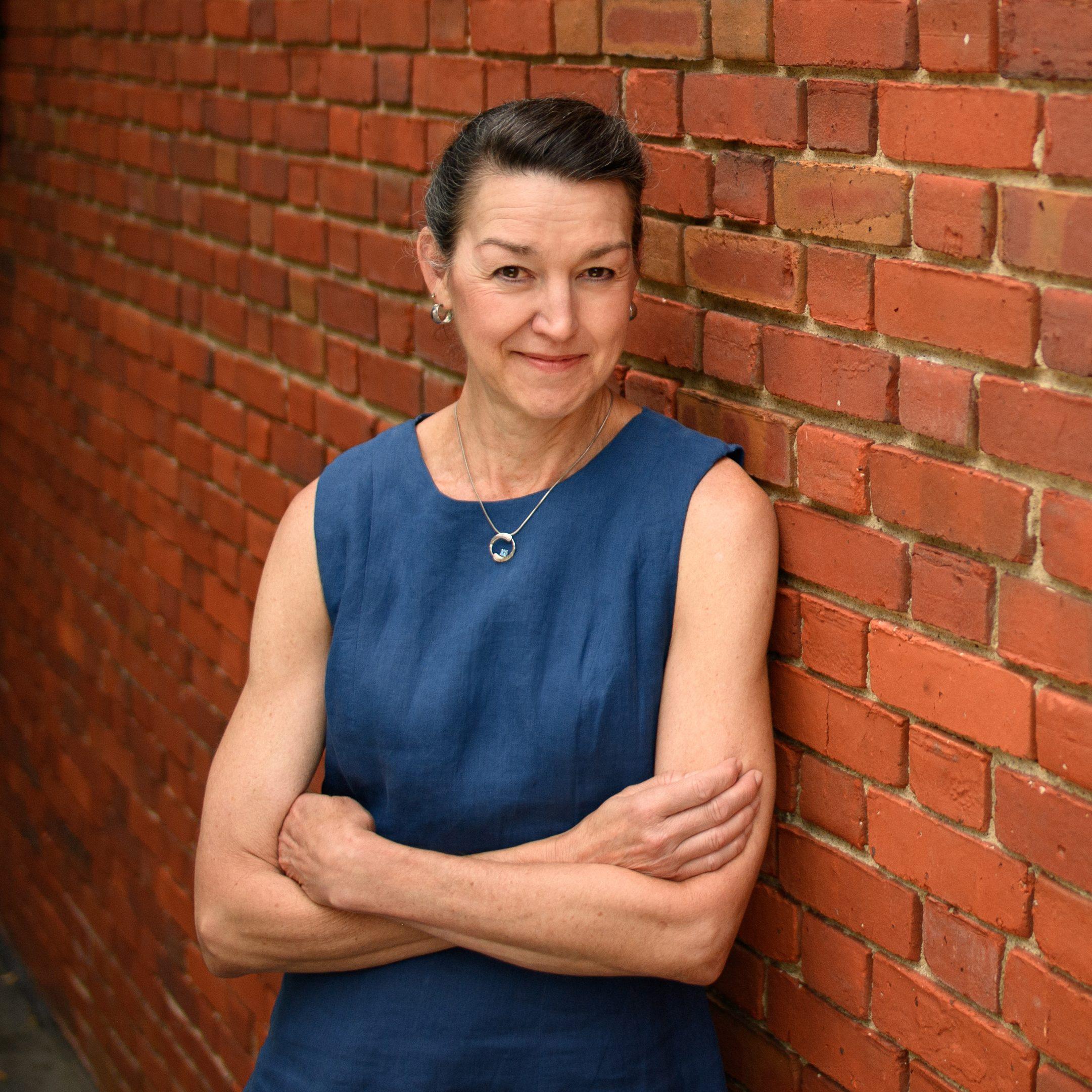A person with short hair in a sleeveless blue dress stands confidently with arms crossed, leaning against a red brick wall. They are smiling and wearing a necklace.