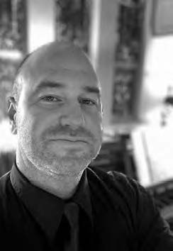 A person taking a selfie in black and white, looking at the camera with a slight smile, wearing a collared shirt and tie. The background is blurred, suggesting an indoor setting with some wall decorations.