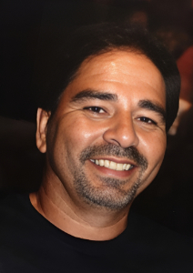 Smiling man with short dark hair and a goatee, wearing a dark shirt, in a dimly lit setting.