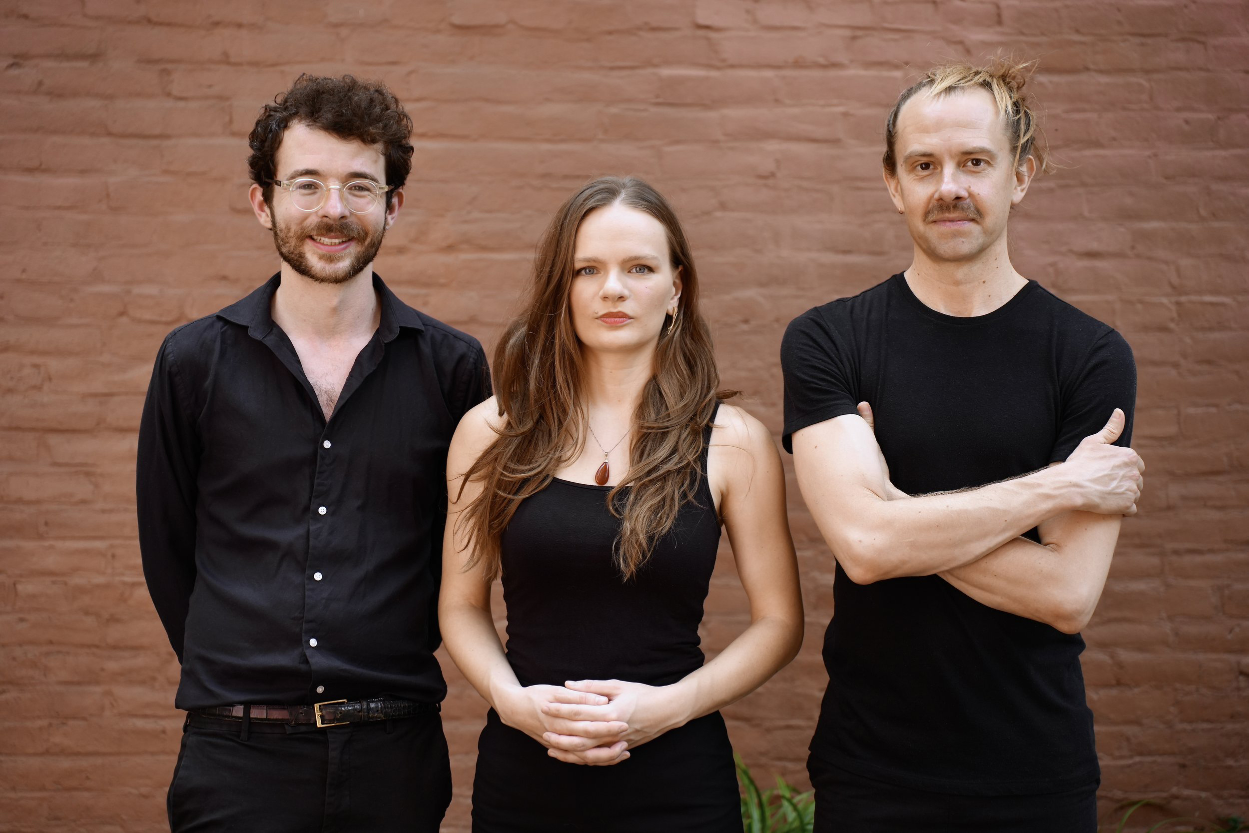 Three people standing against a brick wall. The two men wear black shirts and one has glasses, while the woman in the middle wears a black tank top. They all face the camera with neutral expressions.