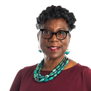 A woman with short curly hair and glasses, wearing a burgundy top and a large turquoise bead necklace and earrings. Shes smiling in front of a plain white background.