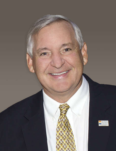 A smiling man with gray hair, wearing a dark suit, white shirt, and patterned tie, stands against a gray background. He has a small pin on his lapel.