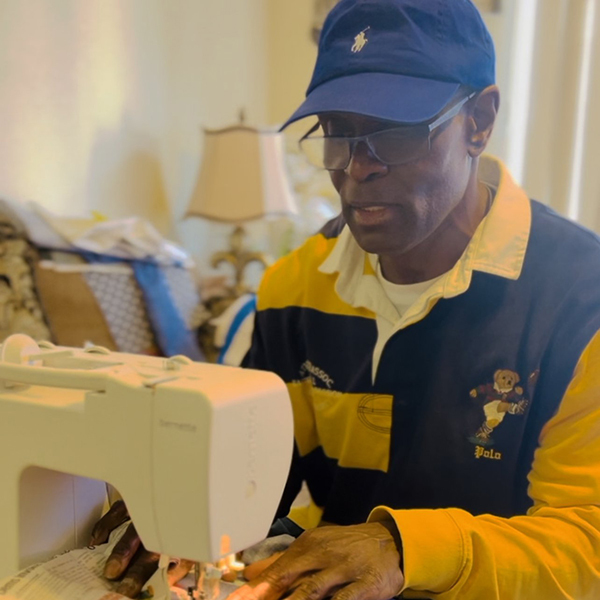 A man wearing a blue cap and striped shirt is focused on sewing at a sewing machine. He is seated in a room with a lamp in the background.