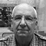 A black and white photo of an older man wearing glasses and a checkered shirt, sitting outdoors. Trees and stone structures are visible in the background.