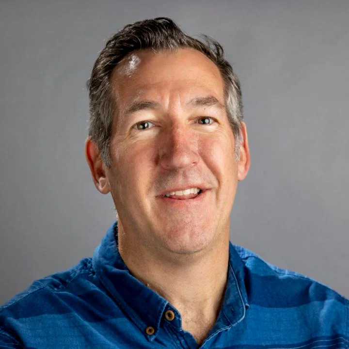 A man with short hair and a small scar on his forehead is smiling at the camera. He is wearing a blue button-up shirt and is set against a plain gray background.