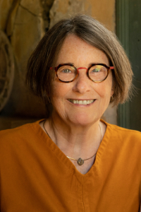 A person with short brown hair and glasses smiles at the camera. They are wearing an orange shirt and a necklace. The background is an indoor setting with warm lighting.