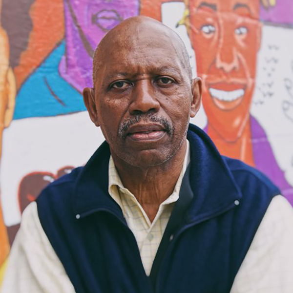 An elderly man with a mustache stands in front of a colorful mural featuring various illustrated faces. He is wearing a dark vest over a light shirt and looks directly at the camera.