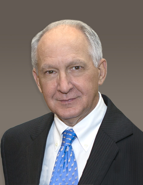 A man with short gray hair is wearing a dark suit jacket, white shirt, and blue tie. He is posed in front of a plain, gradient background.
