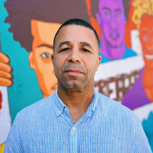A man in a light blue striped shirt stands in front of a colorful mural featuring various cartoon-like faces. The murals vibrant colors include shades of blue, orange, and purple.