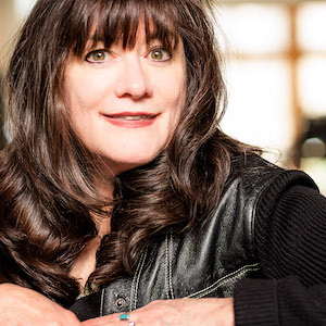 A person with long brown hair is smiling while wearing a black shirt and leather jacket. They are indoors with soft lighting in the background.