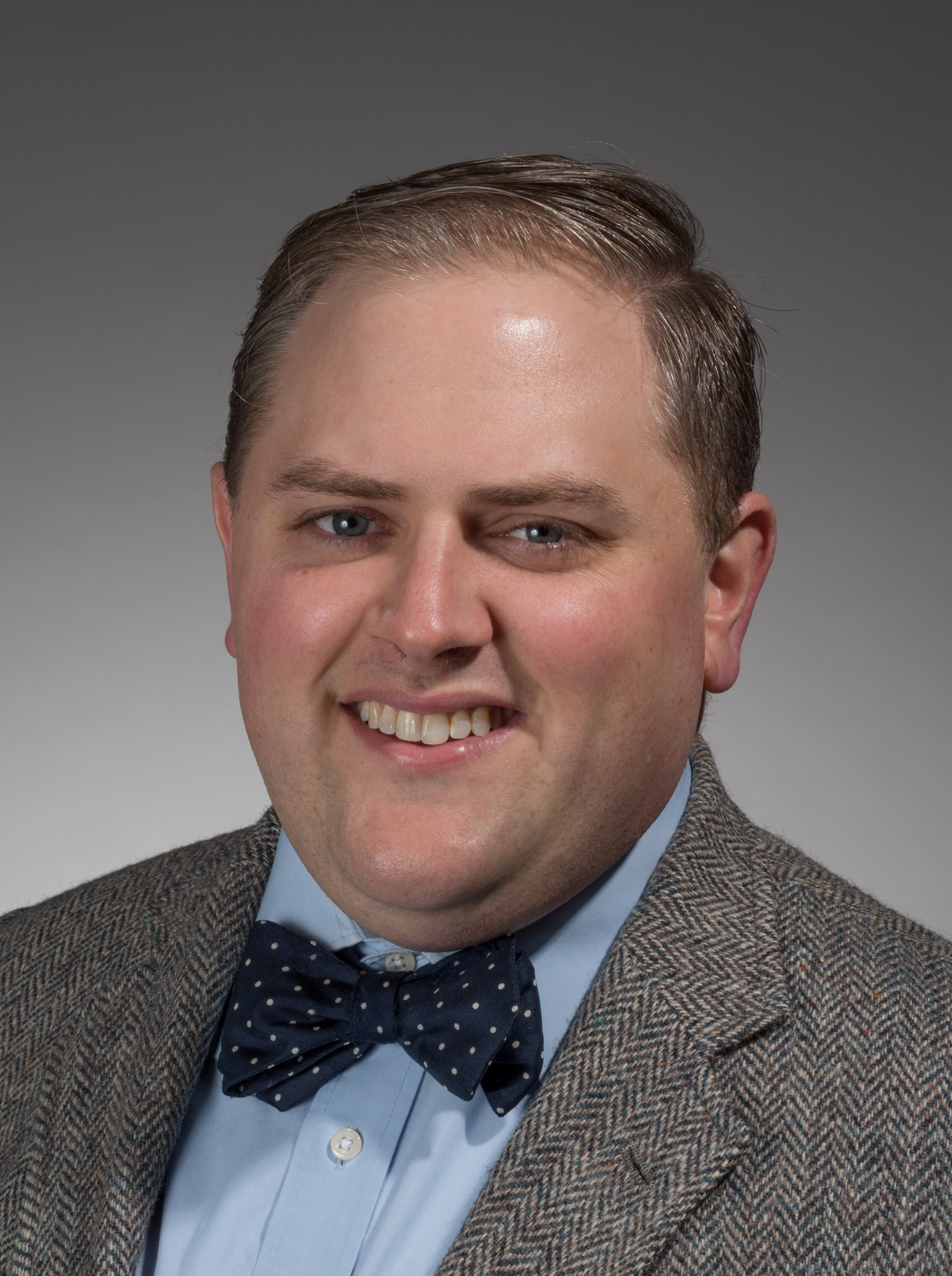 A person with short hair smiles at the camera. They are wearing a herringbone-patterned suit jacket, a light blue shirt, and a navy blue bow tie with white dots. The background is a plain gray gradient.