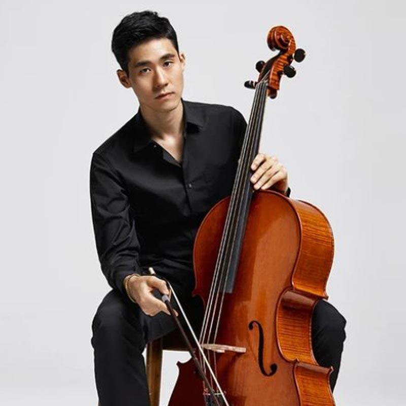 A man in a black shirt sits on a stool holding a cello. The background is plain white, and he appears focused on the camera, with his right hand on the bow and left hand on the fingerboard.