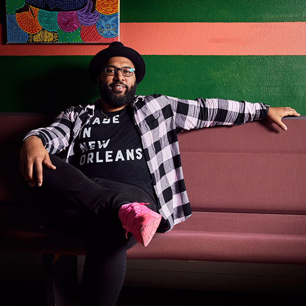 A person wearing a black hat, glasses, and a plaid shirt sits on a maroon bench. They have a T-shirt with Made in New Orleans printed on it and are wearing bright pink shoes. Theres a colorful painting and a green and pink striped wall behind them.