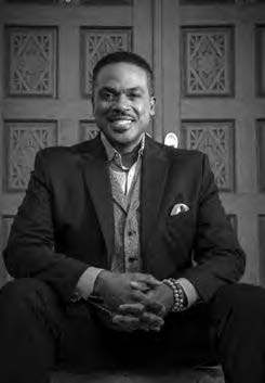 A man in a suit and vest sits smiling in front of a decorative wooden door. His hands are clasped, and he wears a beaded bracelet. The image is in black and white.