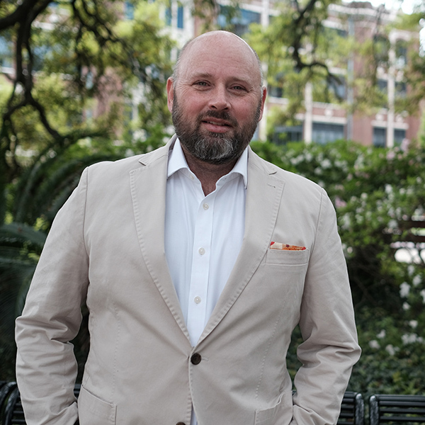 A man with a beard is standing outdoors in front of greenery, wearing a beige suit and white shirt. He has a folded pocket square in his jacket and is smiling slightly towards the camera.