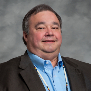 A man wearing a dark suit and a light blue collared shirt, with a multicolored beaded necklace, stands against a gray background. He has short dark hair and is smiling slightly.